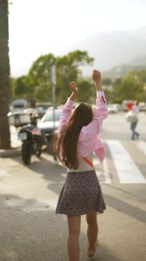 Pembe ceketli, çiçekli etekli şık bir kadın şehirde rahatça dolaşıyor, kaygısız bir hava yayıyor.