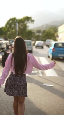 Uzun saçlı bir kadın güneşli bir günde çiçek eteği ve pembe bir bluz giyerek otoparkta yürüyor..