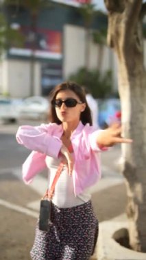 Güneş gözlüklü ve pembe ceketli kaygısız bir kadın şehir merkezinde neşe ve enerji saçarak neşe içinde dans ediyor.