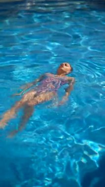 Renkli bir mayo giymiş bir kadın, sakin bir yaz gününde güneşli bir havuzda huzur içinde yüzüyor.