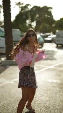 Genç bir kadın güneşli bir günün tadını çıkarıyor, pembe bir ceket ve çiçekli bir etek giyiyor, yürüyor ve eğleniyor..