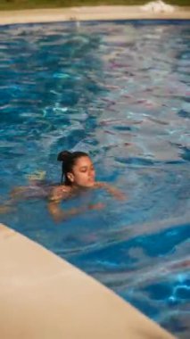 Bir kadın güneşin altında açık mavi bir havuzda yüzerek rahatlama anını yakalıyor.