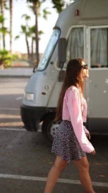 Dışarıda pembe bir gömlek ve çiçekli bir etek giyen genç bir kadın güneşli bir günde palmiye ağaçlarının yanında yürüyor.