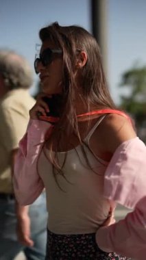 Güneş gözlüklü şık bir kadın, elinde telefon, modaya uygun giysiler içinde dışarıda poz veriyor, kendine güveni ve şık tavırları var.