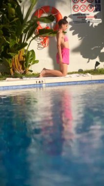 Pembe bikinili bir kadın güneşli bir günde arka planda turuncu bir can simidi olan bir havuzun yanında geriniyor.