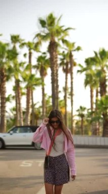 Pembe gömlekli ve çiçekli etekli neşeli bir kadın güneşli bir günde palmiye ağaçlarının altında geziniyor.