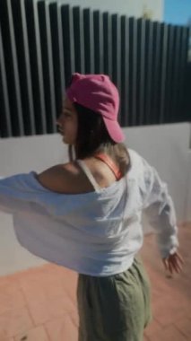 Genç bir kadın açık havada dans etmekten hoşlanır. Pembe bir şapka, beyaz gömlek ve güneşin altında günlük giysiler giyer..