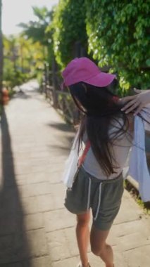 Pembe şapkalı genç bir kız güneşli bir yolda yürüyor, yeşillikler arasında neşe, özgürlük ve canlılık uyandırıyor.