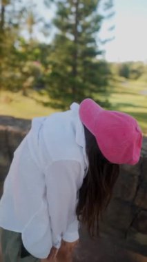 Pembe şapkalı, gri şortlu ve açık havada güneşli gökyüzünün altında dans eden beyaz tişörtlü genç bir kız..