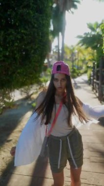 Genç bir kadın parkta, pembe bir şapka takıyor ve gündelik giysiler giyiyor, doğanın ortasında güneşli bir günün tadını çıkarıyor.