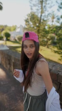 Pembe şapkalı, beyaz ceketli, yeşil ağaçlar ve güneş ışığı olan bir bahçede şort giyen genç bir kadın..