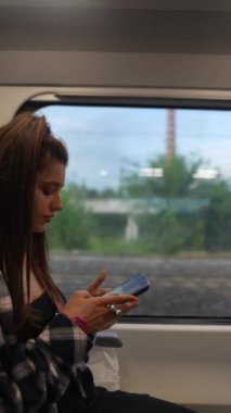 Dışarıda manzarası olan bir trende seyahat eden genç bir kadın, yoğun bir şekilde akıllı telefonuna odaklandı..