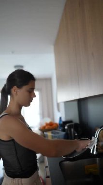 Genç bir kadın, modern bir mutfakta ahşap dolaplar ve modern aletlerle yemek hazırlarken görülüyor.