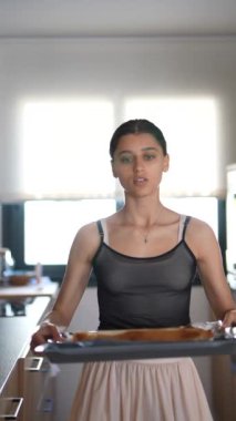 Genç bir kadın modern bir mutfakta taze ekmek pişiriyor, siyah bir üst ve açık etek giyiyor ve tepside çörekler var..