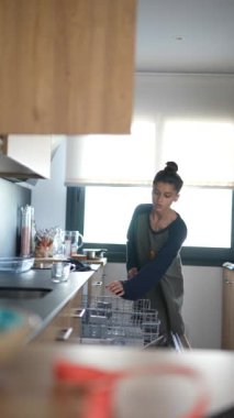 Bir kadın, modern bir mutfakta bulaşık makinesine mutfak gereçleri yüklüyor, gündelik giyinmiş, ev işleriyle meşgul.