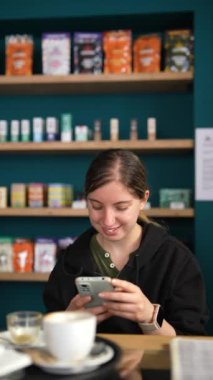 Bir kafede oturan genç bir kadın telefonunu kullanırken kahvenin tadını çıkarıyor. Raflar arkasındakilerle dolu..