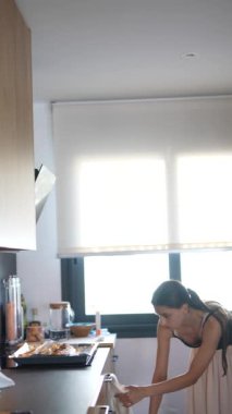 Bir kadın modern bir mutfakta yemek pişiriyor, tepside yemek hazırlıyor ve pencereden güneş ışığı akıyor..