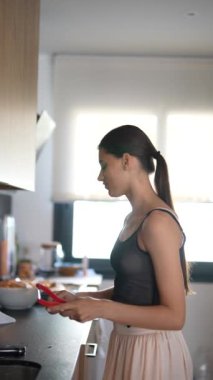 Genç bir kadın modern bir mutfakta tasvir ediliyor, yandan görülüyor, doğal ışık altında kahvaltı hazırlıyor.