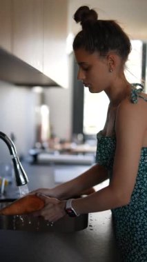 Genç bir kadın mutfaktaki lavaboda tatlı patatesleri yıkıyor. Çiçekli bir elbise giyiyor..