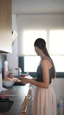 Genç bir kadın modern bir mutfağa odaklanmış, özenle yemek pişiriyor ve hazırlıyor.