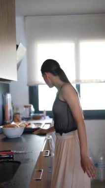 Modern bir mutfakta, genç bir kadın kahvaltı hazırlar, etrafı toplar, yemek düzenler ve tezgahı siler.