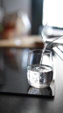 Su, bir sürahiden siyah bir tezgahın üzerindeki bir bardağa nazikçe dökülür, saflığı ve yenilenmeyi simgeler.