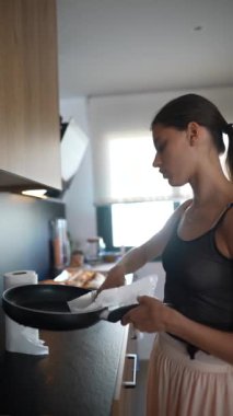 Genç bir kadın parlak, modern bir mutfağa kızartma tavasını temizleyip kaldırıyor..