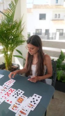 Gündelik kıyafetli genç bir kadın, güneşli bir terasta, yemyeşil bir bahçeyle mutlu bir şekilde kağıt oynuyor.
