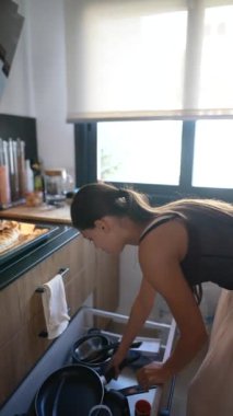 Parlak, çağdaş bir mutfakta, bir kadın bulaşıkları yıkar, temizliği ve ev yapımını ön plana çıkarır.