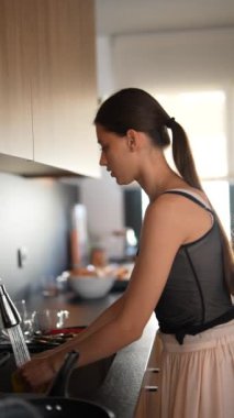 Gündüz vakti modern bir mutfakta bulaşık yıkayan, siyah bluz ve açık etek giyen bir kadının arka planı..