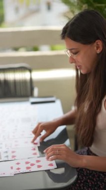 Odaklanmış genç bir kadın güneşli bir günde teras masasında kağıt oynayıp dışarıda eğleniyor.