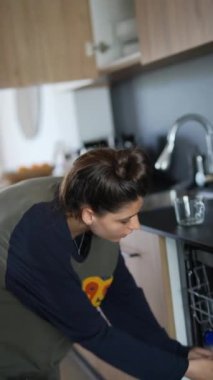 Modern bir mutfakta bir kadın bulaşık makinesine bulaşık dolduruyor, ev işlerini yaparken düzenli bir yaşam alanı sağlıyor.