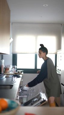 Günlük kıyafetleriyle mutfağını toplayan, bulaşık makinesini dolduran ve verimli bir yer için dolaplar düzenleyen bir kadın.