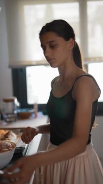 Genç bir kadın modern bir mutfakta yemek pişiriyor, gri atlet ve açık pembe etek giyiyor, orta yemek hazırlıyor.