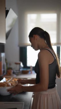 Modern bir mutfakta, at kuyruklu uzun saçlı genç bir kadın odaklanır ve konsantrasyonuyla yemek hazırlar.