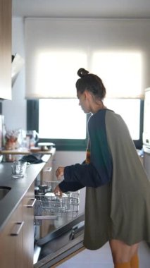 Bir kadın modern mutfağını bulaşık makinesine bardakları istifleyerek ve eşyaları dolaplara yerleştirerek düzenler..