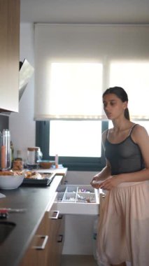 Modern bir mutfakta, çekmecede çatal bıçak düzenleyen ve kahvaltı hazırlayan genç bir kadın. Parlak, rahat bir ortam..