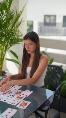 Gözlük takan ve beyaz bluzlu genç bir kadın dışarıdaki cam bir masada, bitkilerle solitaire oynuyor..