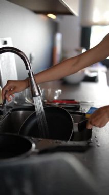 Modern bir mutfakta, bir kadın lavabonun altındaki bulaşıkları yıkıyor, başlıca amacı kızartma tavasını fırçalamak.