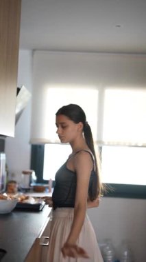 Modern bir mutfakta, gündelik giysiler içinde genç bir kadın fırının yanında yemek pişirir, rahat ve davetkar bir ortam yaratır.