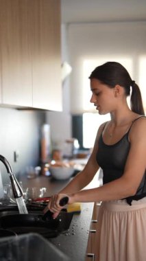 Bir kadın, modern bir mutfakta bulaşıkları yıkıyor. Pencereden akan güneş ışığıyla aydınlatılıyor.