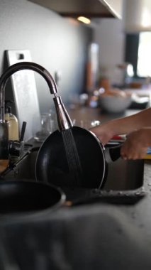 Mutfak lavabosunda bulaşık yıkayan, akan su ve süngerle siyah bir tavaya odaklanan kişinin yakın plan fotoğrafı..