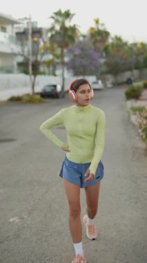 Canlı genç bir kadın şehirde koşuyor, güneşli bir günde pembe kulaklıklarıyla müziğin tadını çıkarıyor.