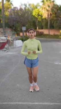 Genç bir kadın açık havada koşusunun keyfini çıkarırken yeşil bir bluz, mavi şort ve kulaklık giyer..