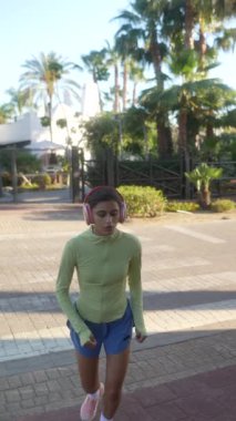 Genç bir kadın tropikal şehir ortamında koşuyor, kulaklık takıyor, yeşil bluz ve mavi şort giyiyor..