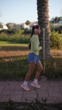 Canlı giyinmiş genç bir kadın güneşli bir günde şehir parkında koşuyor ve müzik dinliyor.