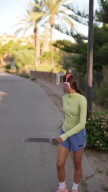Güneşli bir günde genç bir kadın, palmiye ağaçlarıyla çevrili bir caddede kulaklık ve spor kıyafetleriyle koşuyor.