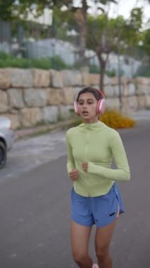 Genç bir kadın pembe kulaklıklarla koşuyor, yeşil bluzlu ve mavi şortlu, açık havada spor yapıyor..