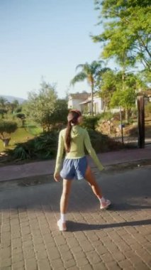 Yeşil bluzlu ve mavi şortlu genç bir kadın güneşli bir günde açık havada egzersiz yapıyor ve kulaklıkla müzik dinliyor.