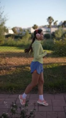 Bir kadın güneşli bir günde dışarıda koşuyor, kulaklık takıyor ve spor yaparken spor kıyafetlerini giyiyor.